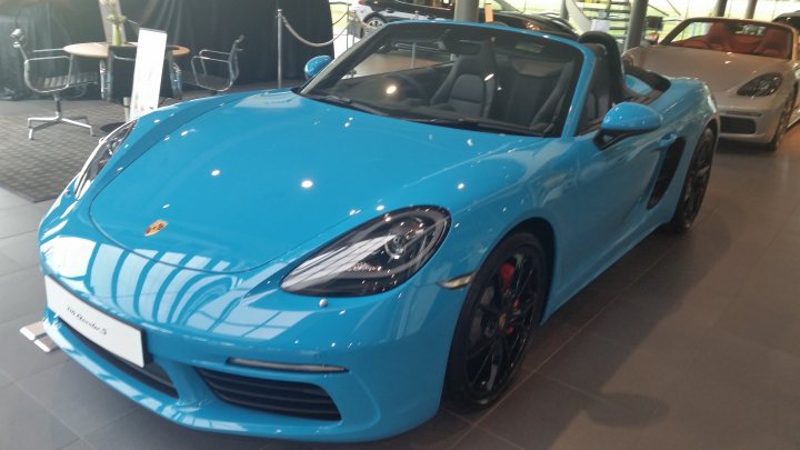 718 Cayman Spec & Colours- what have you gone for? - Page 74 - Boxster/Cayman - PistonHeads - The image shows a bright blue sports car parked indoors, likely on a showroom floor or at a car dealership. The vehicle is a Porsche 718 Cayman, as indicated by the logo on the hood. The interior area surrounding the car features a modern architectural design with floor-to-ceiling windows, smooth surfaces, and a minimalist aesthetic. The lighting in the room highlights the glossy finish and sleek lines of the car. The vehicle's vibrant blue color stands out against the more neutral tones of the showroom space.