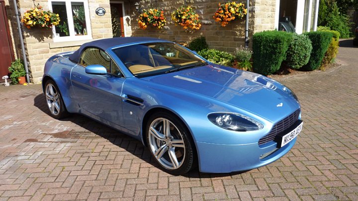 Detailing Experience Pistonheads - The image shows a blue luxury roadster parked on a brick driveway. It has a sleek design with a convertible top, visible grille, and halogen headlights. The car is positioned on the wheel, showing off its alloy wheels. In the background, there's a brick house with a small garden area. There are decorative planters with flowers and shrubs around, and a door on the left side of the image. There is no text visible in the image.