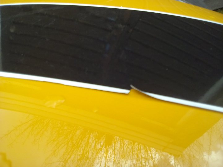 F56 bonnet stripes lifting / peeling - Page 1 - New MINIs - PistonHeads - The image focuses on a section of a yellow car. The most striking feature is a large black spot that stretches across the image, which is likely the reflection of the sky or surrounding environment. Above the black area, there is a small portion of the sky or another environment, but it is mostly obscured by the black spot. The car's paint appears matte with some slight reflections near the edges. The bottom of the image reveals a hint of the ground, suggesting the car is parked on a tarmac or similar surface. The image composition emphasizes the stark contrast between the yellow of the car and the black of the reflected element.