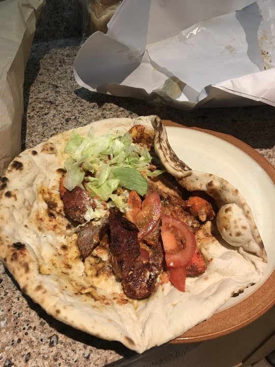 A slice of pizza sitting on top of a white plate - The image presents a close-up view of a delectable meal placed on a table. A large pita, possibly filled with meat and vegetables, is the central focus of the image. It's garnished with a generous serving of lettuce, adding a pop of green to the dish. The pita is served on a white plate, which contrasts nicely with the brown color of the table. 

In the background, there are two sandwiches and a pizza, suggesting a variety of food options available in this setting. The entire scene appears to be set indoors, possibly in a casual dining environment or at someone's home. The food items are arranged neatly on the table, ready to be enjoyed.