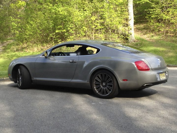 I’ve put my football boots on & bought a Bentley GT Speed - Page 1 - Rolls Royce & Bentley - PistonHeads - The image features a gray metallic car parked on a paved parking area. The car has a fastback design, characterized by its sloping back window and rear door. The vehicle is positioned facing the right side of the image, revealing the front and right side profile. The backdrop is a serene outdoor setting with green vegetation and a clear sky.