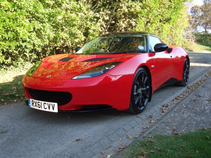 Pistonheads - The image features a bright red sports car parked on a roadside. The car is a convertible with a soft top, as indicated by the presence of a top behind the rear windshield that is not raised. It has a sleek design with a low profile and large, black wheels. The bodywork of the car is somewhat reflective, suggesting a glossy finish, and there are badges visible on the front and rear, but not enough details to discern the make or model. The roadside itself is not overly busy and is bordered by grass on one side and a hedge on the other, suggesting a setting outside of urban areas.