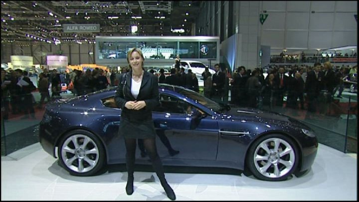 Pistonheads Axed Gear - In the image, a woman stands confidently in a showroom, her arms crossed in a symmetrical pose. She is positioned to the side, slightly ahead of the blue car she's highlighting. The car, characterized by its silver wheels, takes center stage in the image. The showroom itself is filled with onlookers, adding to the atmosphere of the event. Large windows punctuate the background, allowing natural light to flood the space.