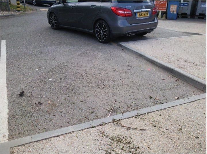 The BAD PARKING thread [vol3] - Page 463 - General Gassing - PistonHeads - The image depicts a small, hatchback car parked on the side of a street. The car is gray with a noticeable plot number visible on its license plate. The street appears to be made of layered gravel, giving it a textured look. There is litter scattered on the floor near the sidewalk, indicating some level of neglect or a recent refuse disposal. In the distance, other parked cars can be seen, indicating that this might be a public parking area or a street with on-street parking.