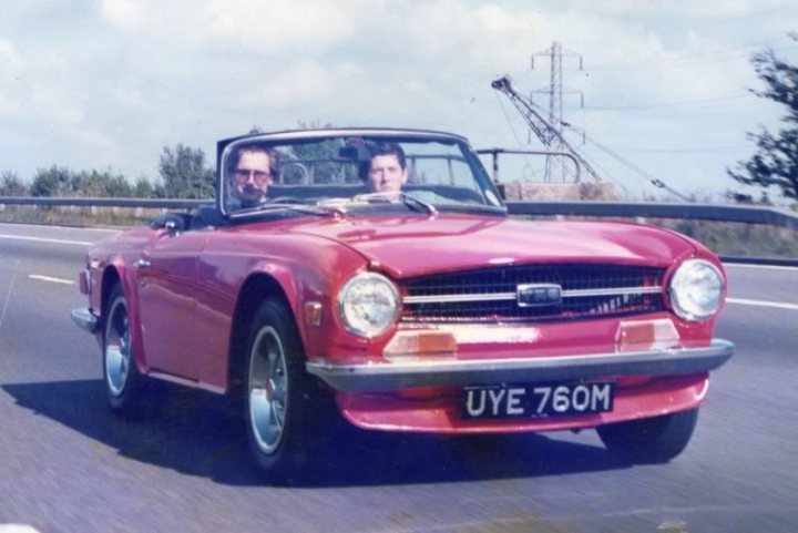 Tr6 wow! - Page 1 - Triumph - PistonHeads - The image captures a dynamic scene of a red convertible car with two individuals driving on a highway. The vehicle is in motion with the convertible top down, allowing the passengers to fully enjoy the clear day. In addition to the car, there is a clarifying sign with the letters "UEY 760M" in the background, possibly indicating a location or route. The highway itself appears to be in a rural setting, given the visible trees along the side and a tall structure looming over the landscape. The image conveys a sense of adventure and leisure, as well as a sense of speed, emphasized by the blurred background.