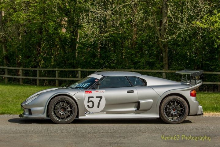 Cars in the park newby hall  - Page 1 - Noble - PistonHeads - The image showcases a sleek, gray sports car parked in a forested area. Its metallic sheen is prominently reflected on its body, adding to its allure. A distinctive number, "57", is prominently displayed on the hood, suggesting it might be a race car. Another number, "56", is written on the door, matching in style with the car's overall theme. The vehicle is parked on a gravel road, with a fence and trees visible in the background, creating a serene and natural setting for the car.