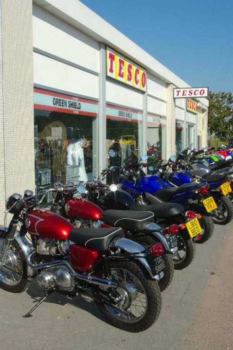 breakfast club sunday october 2 - Page 1 - Goodwood Events - PistonHeads - The image shows a line of motorcycles, or mopeds, parked along the sides of a street. The motorcycles vary in color, including blue, red, and green. They are parked in front of a building that has a display of clothing in its window. The store sign above the building reads "Tesco." The setting appears to be an urban area, possibly on a sunny day given the shadows on the ground. The motorcycles seem to be parked securely, and the variety could suggest a custom shop or a dealership.