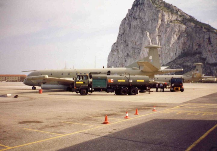 Pistonheads - This image depicts a scene on an airport tarmac. The central focus is a large aircraft, painted in a camouflage pattern of white, green, and gray, possibly an army transport plane, dwarfed by a massive rock formation in the backdrop, reminiscent of rocks at Blackresent Point in Gibraltar. The plane has a prominent fold-out ramp, indicating it's designed for quick loading and unloading. Surrounding the plane are several smaller vehicles, including a green utility vehicle, likely servicing or supporting the aircraft's operations. The tarmac is marked with yellow lines and orange cones, indicating a well-organized airport area. The surrounding environment suggests a military or government-owned airport.