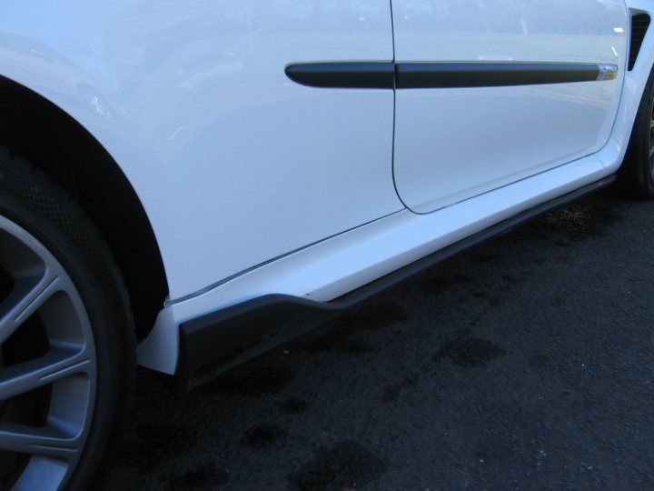 A car parked in front of a parking meter - Pistonheads - This image shows a close-up view of the front of a white car, focusing on the side skirt and wheel. The car is parked, and the surface underneath it appears to be a road or parking area. The lighting suggests it's either a cloudy day or under an overhang. There is a reflection of light on the car's side, indicating some ambient light. The image provides a view of the car's design detailing, specifically the shadow cast by the side skirt.