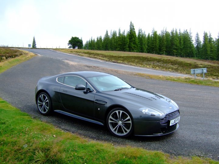 Who has a V12 Vantage? - Page 19 - Aston Martin - PistonHeads - The image shows a sleek, gray sports car parked on the side of a winding country road. The car, a two-door luxury model, has large, silver-rimmed wheels and features the distinctive badge of the Bentley Motors on the front. Its reflections indicate that it's a sunny day. The road is flanked by lush green grass and dense forest of tall trees, providing a serene and natural backdrop. There is no text in the image that is legible. The car appears to be in pristine condition with a glossy finish, possibly maintained by the car's owner.