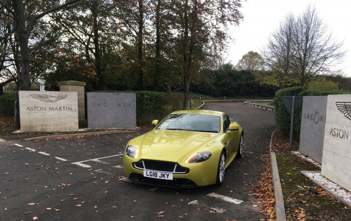 So what have you done with your Aston today? (Vol. 2) - Page 115 - Aston Martin - PistonHeads UK - The image depicts a scene with a light green sports car parked in front of a sign that reads "Aston Martin." The car is sleek and modern, suggesting it's a high-end vehicle. The setting appears to be an entrance or parking area, as there's a driveway leading up to the car. The environment around the car includes trees with autumn leaves, indicating the season might be fall or early winter. There are no people visible in the image, and the focus is solely on the car and its immediate surroundings.