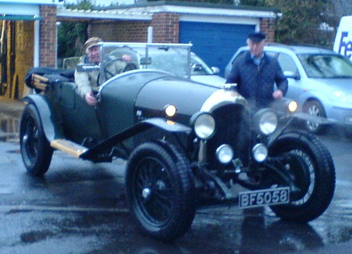 Tell me about this Bentley, please - Page 1 - Rolls Royce & Bentley - PistonHeads - This image captures a moment involving a vintage vehicle, specifically an old-fashioned car characterized by its round headlights and black tires. Two men are present in the scene, with one man, dressed in dark attire and a hat, appearing to ensure the car is safe for its journey. The other man, presumably the driver, is positioned behind the wheel. The setting appears to be a street, with a house and a garage visible in the background. The sky overhead presents a gloomy atmosphere, possibly indicating an overcast day or an approaching storm.