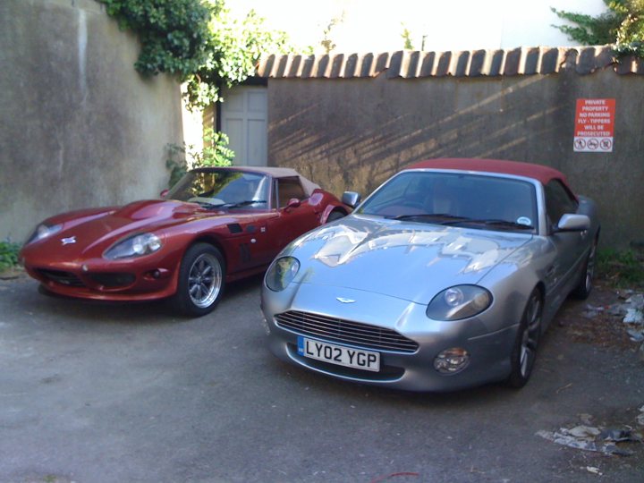 The Proper Colour for an Aston thread - silver, greys, black - Page 1 - Aston Martin - PistonHeads - In the image, there are two sports cars parked side by side on a brown, asphalt surface. The car on the left is a vibrant red, while the one on the right is a sleek silver. Both cars have a convertible top style, suggesting they are suitable for open-air driving. The background showcases a brick wall with a lobby that has a futuristic design. Also in the background, there are traffic cones and a sign standing out contrastingly against the wall.