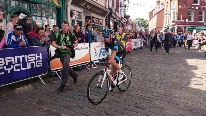 Pistonheads - The image captures a moment in a bicycle race. A cyclist, dressed in a black uniform with white accents and a blue visor, is actively pedaling down a downhill course with brick paving. The cyclist is crouched low over the bike, pointing towards the right side of the image, demonstrating a meal-on-wheels approach to minimize air resistance. 

The race is taking place on a narrow stone street closed off for the event. A crowd of spectators, visible in the background, watches the race, adding a sense of excitement and anticipation to the scene. Their presence suggests a public event or tournament of some sort. 

The ambiance of the scene is further enhanced by the presence of shops and a restaurant in the background, indicating an urban setting. The race seems to be well-organized, with people in reflective vests guiding the racecourses and spectators respecting the barriers set up for safety. The overall image paints a vivid picture of a dynamic, competitive bicycle racing event.