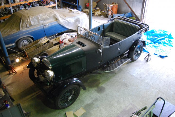 Pistonheads - This image captures an indoor scene where an old, green antique car is parked, appearing to be in a workshop or garage. The car is the main focus of the image, its vintage charm accentuated with headlamps that add a touch of classic elegance. Behind the car, you can see a wall strewn with various objects, suggesting a working or repair space. A wooden door is visible beside the car, possibly leading to another area of the building. The setting is well-lit, allowing details of both the car and the workshop to be clearly seen.