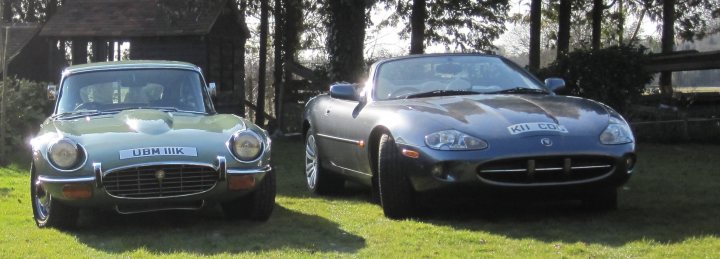 A car is parked in a field next to a car - Pistonheads - The image depicts two classic sports cars parked next to each other outdoors. The car on the left is a silver, with black text indicating it's a Lotus Seven, a well-known British sports car. The car on the right is a deep blue and is described as a "kit car," which typically refers to a car that is custom-assembled from a kit of parts. Both cars have the skirt on their body, adding an element of speed and style. The background suggests a quiet, park-like setting with trees and some sort of structure, which might be a utility building or similar. The presence of two such cars side by side hints at a shared interest in automobiles or possibly a car show or meetup.