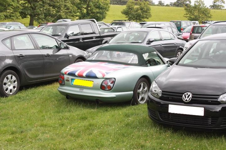 Pistonheads - The image shows a parking lot filled with various cars and two Volkswagen Touaregs. The cars are parked haphazardly, indicating a public parking area. A conspicuous feature is a small, silver-colored sports car with an iconic British flag on its hood, sung res, and its registration tag is yellow. The rest of the cars are more typical and black-colored, suggesting a variety of vehicles. In the background, there appears to be a grassy area, and the sky is visible, indicating an open-air setting. The style of the image is a standard photograph, taken in natural light.