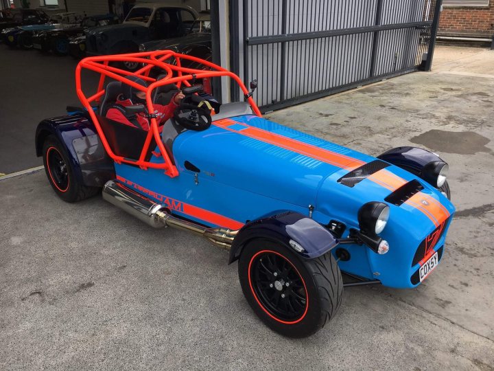 New bespoke 420R - Page 1 - Caterham - PistonHeads - The image showcases a vintage car with an orange roll cage running through its center, featuring an open top. The vehicle is parked on an empty asphalt surface next to a metal fence. The same color is used throughout the car's body, with the trimmings and trim stripe matching the roll cage. The license plate of the car is visible in the lower foreground, adding a personal touch to this distinctive vehicle.