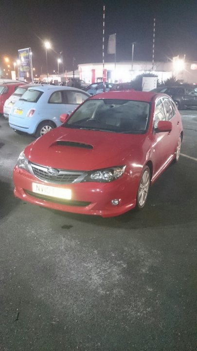 A red car is parked on the side of the road - Pistonheads - The image shows a dark night scene in a parking lot. A red car, which appears to be a Ford Mustang, is parked prominently, occupying most of the frame. Its front lights are ON, illuminating the immediate surroundings. The car has a distinctive sleek design, typical of sports cars. Other cars of various colors and models are visible in the background, suggesting this is a busy area. The gloom of the night, combined with the artificial lighting, creates a stark contrast with the red car, making it the standout feature of the image.