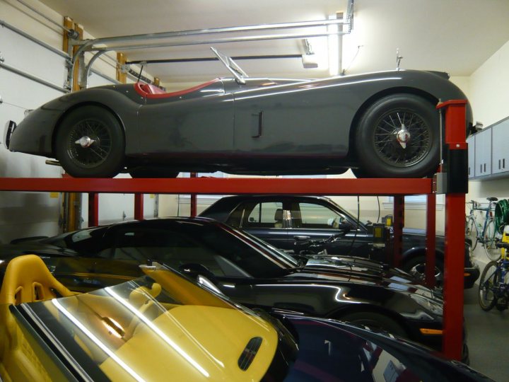 Pistonheads Open Season Jaguar - The image shows an indoor space, possibly a garage or storage facility, with several vintage cars. The cars are parked in what seems to be an organized manner. In the foreground, there is a black car with a chrome gas tank. Above the black car, on a red rack, is a vintage silver car. Additionally, a black convertible with a red interior is elevated on a red rack. The setting looks industrial and utilitarian, with metal poles and ceiling fixtures.