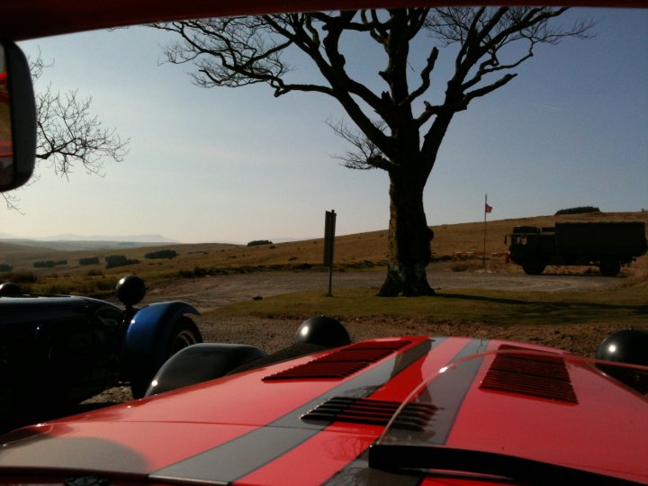 Pistonheads Wales Blat - The image, captured from the perspective of a car windshield, offers a unique view of a military-style humvee parked in a dirt lot next to a grassy area. A striking red car featuring a black stripe down the middle is facing us, giving us a close-up view of its hood. Beyond the vehicles, a large tree stands prominently in the scene, overlooking a field where a flag is visible, adding to the outdoor, possibly military, setting. The sky overhead is clear, suggesting bright, possibly sunny, weather conditions.