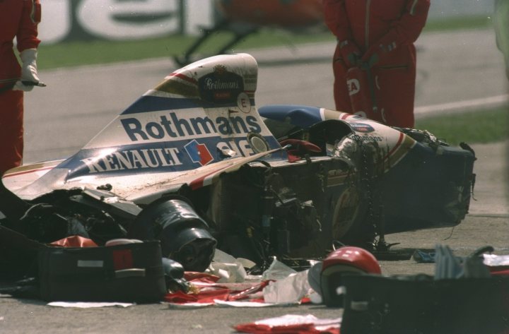 Slightly different footage of Senna's crash... - Page 5 - Formula 1 - PistonHeads - The image shows a heavily damaged race car on a track. It's a Rothmans Haas CART race car with the number 5 and the word "renault" visible. The car's bodywork is severely crushed, and the front end is almost completely detached. Safety equipment, such as a helmet, is scattered around the track, indicating a violent accident. Two people in red suits are standing next to the wreckage, possibly assessing the situation.