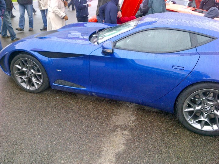 Supercar Sunday pictures, post them up! - Page 1 - Goodwood Events - PistonHeads - The image shows a sleek, blue sports car parked amidst a crowd as part of what appears to be a motor vehicle show. The vehicle's design suggests it's likely a high-performance model, with aerodynamic features such as a rear wing. The hood is propped open, revealing the engine and potentially attracting attention from the attendees. These people, seen in the background, are dressed in various casual outfits and seem to be enjoying the event, adding to the lively atmosphere of the show.