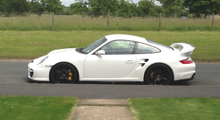 996 GT2 - Is this a mad price or what? - Page 3 - Porsche General - PistonHeads - The image showcases a white Porsche Cayman sports car, prominently displayed on a grassy area bordering a road. The car is positioned at an angle, allowing a clear view of its sleek design and the convertible top folded down, enhancing its sporty aesthetics. Its side profile reveals razor-sharp wheel arches and black alloy wheels, contributing to its luxury and performance image. The surrounding environment, with its neatly trimmed grass, contrasts with the car's sleek and aggressive design. There appears to be a small footpath or pavement to the right of the car, possibly leading towards a parking area or residential property. The overall scene conveys a sense of luxury and leisure, typical of a sunny day with a sports car.