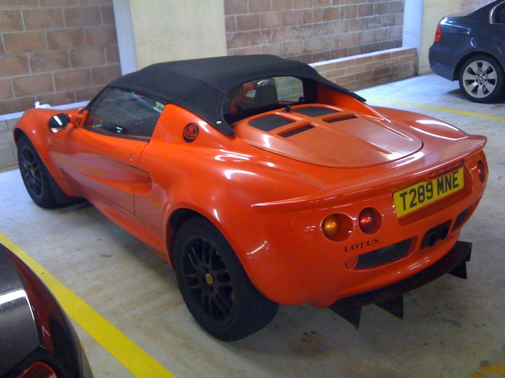 Pistonheads Thinking Selling - The image showcases a striking, vivid orange Lotus sports car, parked in a garage. The car is characterized by a glossy exterior and a sleek black interior. It features a black soft top roof, which is down in this instance. Its license plate proudly displays "Lotus." The car is positioned against the backdrop of a sturdy brick wall, which punctuates the otherwise smooth surface.