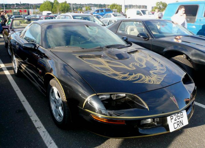 Pistonheads - The image is a photograph featuring a black sports car with a gold and red flame design on the hood, which is parked in a parking lot. The vehicle displays a model-specific license plate that reads "P228 CRN." The car has yellow headlight accents and modified wheel rims, suggesting aesthetic modifications. In the background, there are other vehicles, indicating this is a public parking area. The parking lot itself appears to be in an outdoor area and there are individuals visible in the distance.