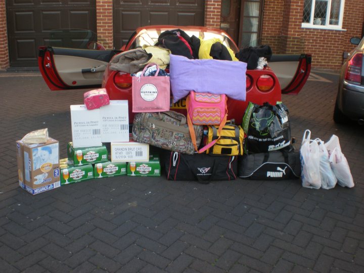 What an all rounder the Vette is - Page 1 - Corvettes - PistonHeads - The image shows a red sports car loaded with various items. There are personal belongings including bags with vibrant patterns, pink shopping bags, and boxes of what appears to be Heineken beer. The fuel door of the car is visible, revealing the trademark lifestyle bag. The items are placed on the ground behind the car, suggesting the vehicle may be moving or an on-the-spot loading of luggage. The setting is an outdoor parking lot with a building in the background.