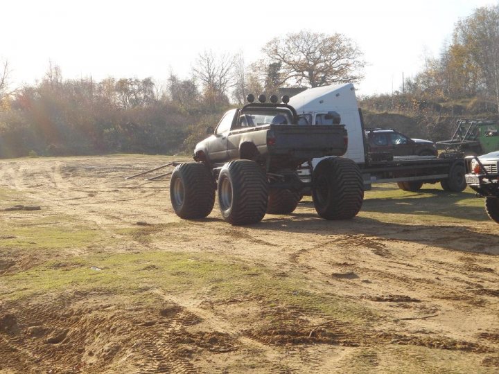 Pistonheads - The image captures a scene on a dirt lot with a large, old utility truck as the prominent vehicle. The truck is heavily customized, featuring adhesive stripes and large tires on its flatbed trailer, which stands on six wheels. The background reveals other vehicles and a barren landscape, hinting at a possible industrial or construction site setting. The lighting and shadows suggest it is daytime, with the truck positioned on a sloped terrain under the open sky.