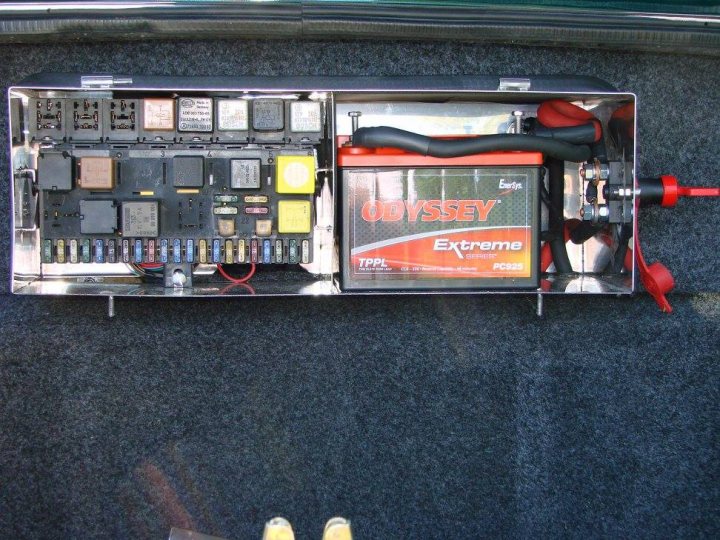 Battery and Fusebox relocation - advice please - Page 1 - Griffith - PistonHeads - The image shows an open car trunk. Within it, there is an arrangement of dashboard-style gauges and odds and ends. Prominently visible is the company name "Odyssey" written on one of the components, suggesting that the battery or power source is from Odyssey. There is also a cord or hose connected to a red adaptor of some sort. The overall setting appears to be a garage or open-air vehicle maintenance and storage area due to the presence of the open trunk and the array of electronics and components.