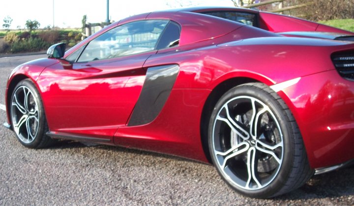 A red and black car with a red umbrella - Pistonheads - The image showcases a striking red sports car parked on a street, with a focus on its large black wheels. The vehicle's aerodynamic design, with a low and sleek profile, suggests it is built for speed and performance. The setting appears to be a road lined with trees and a lamppost, suggesting an urban or suburban environment. The lighting suggests it might be late afternoon or early evening.