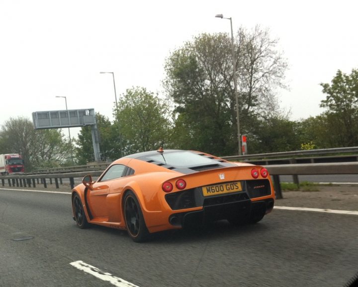 Supercars spotted, some rarities (Vol 4) - Page 300 - General Gassing - PistonHeads - This image captures a high-speed scene on a highway. The main subject is a bright orange sports car, leaning slightly to one side, indicating motion and speed. The car is adorned with what appears to be a regal griffin emblem, adding to its luxurious appearance. The highway itself is lined with trees and a guardrail, providing a sense of depth and scale to the image. In the distance, other highway-related objects such as traffic lights and signs are visible, further emphasizing the high-speed nature of the scene.