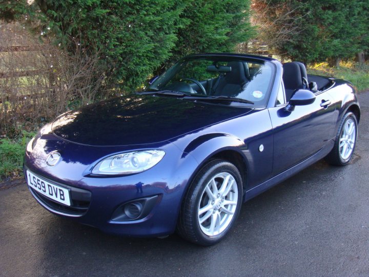 MX5 Mk3 or Mk3.5 - Page 1 - Mazda MX5/Eunos/Miata - PistonHeads - This image features a striking black two-door convertible parked on the side of a road. The car, which is a FyNNage, has a modern design with alloy rims and a convertible top. The setting appears to be a rural or suburban area with verdant shrubbery in the background, suggesting a tranquil environment. The sun seems to be shining, casting a bright light on the car, making its color and features more prominent.