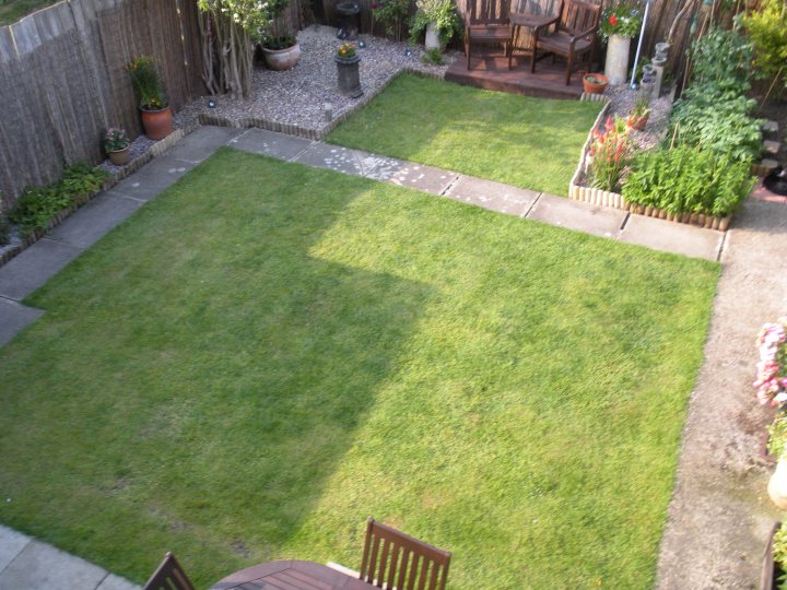 Pistonheads - This image showcases an outdoor space that seems to be a combination of a courtyard and a garden. The area is divided by a path leading to another section, and it's covered in freshly cut grass. Surrounding the grass are planted beds filled with vibrant flowers and shrubs. To the left and right of the image are wooden seating areas, suggesting this space might be used for relaxation and enjoyment of the natural surroundings. Around the area, white and green potted plants can be seen, adding to the serene and well-maintained atmosphere.