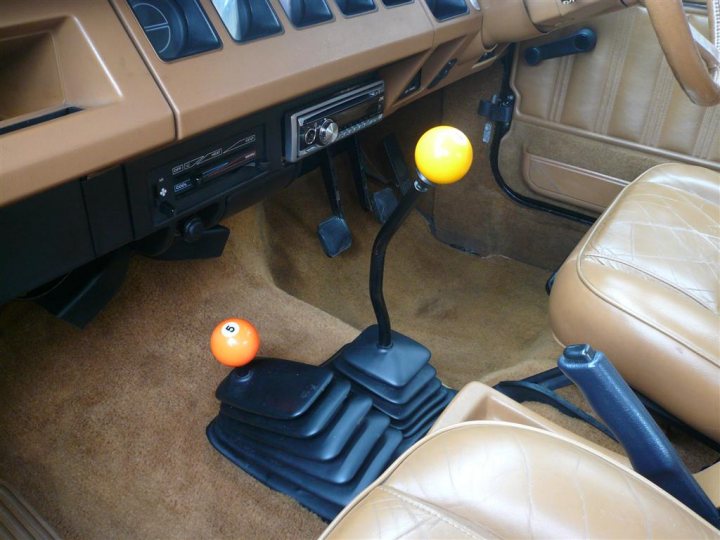 Americana Naff Pistonheads Suitable - This image captures the interior of a vehicle with a distinctive setup. The central focus is on a custom gear shifter, which is located in the center console of the vehicle. It's colored black and appears to be part of a personalized modifier kit. This shifter is flanked on either side by two orange balls, which seem to serve as cup holders, adding a splash of color to the otherwise neutral interior. The vehicle features cream or light-colored upholstery, and the overall impression is of a well-kept and unique vehicle interior.