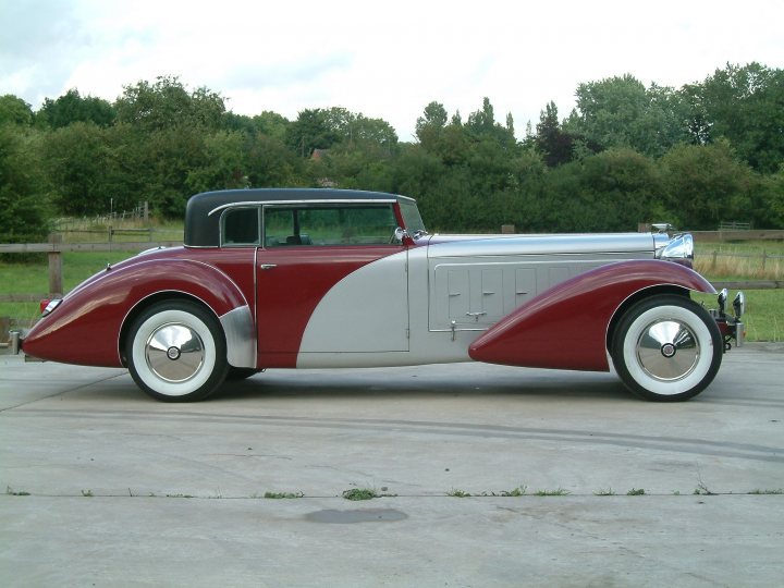 Pistonheads Style Kit - The image shows a vintage motorcar with a classic design, featuring a combination of red and white bodywork. The car has a streamlined form with a sloped front and a flowing, jagged side design. It is parked on an arid, ground surface with sparse patches of grass, and there is a wooden fence and trees in the background, suggesting the location might be rural or on the outskirts of a town or city. The vehicle's presence here adds a touch of antiquity to the otherwise ordinary environment.