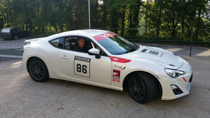 A car parked next to a parking meter - Pistonheads - The image shows a white racing car parked in a lot, with a person in the driver's seat. The vehicle is associated with the Toyota brand, as indicated by the number 86 on the side. There is a red sponsorship sticker on the door. The car's sleek design, dynamic lines, and alloy wheels suggest it's built for speed and possibly used for racing or high-performance driving. The setting appears to be a typical parking area with greenery in the background.