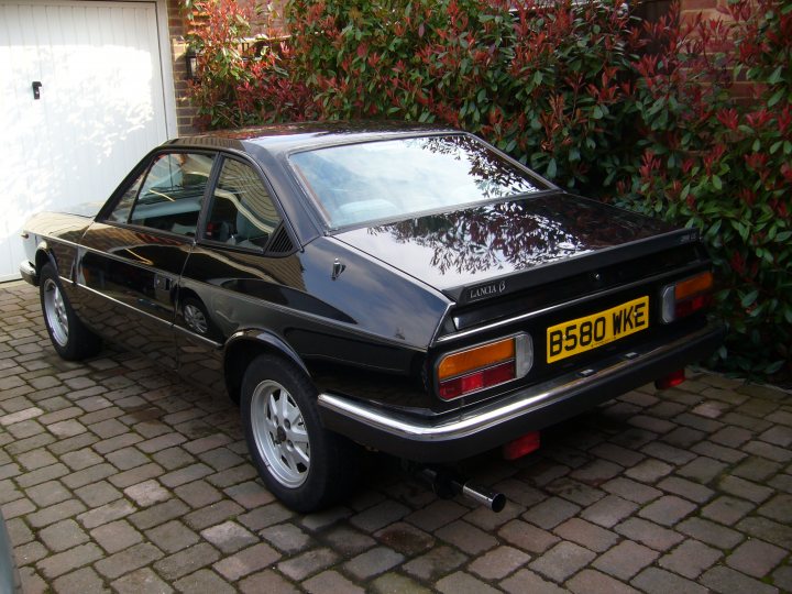 Pistonheads - The image is a photograph of a small, dark-colored car parked on a brick driveway next to a closed garage. The vehicle has a noticeable yellow license plate at the back, and the make and model appear to be from a European country. The car features a spoiler on the trunk, and there are also what seem to be two exhaust pipes visible below the rear of the car. The surroundings include lush green bushes, suggesting a well-maintained garden or outdoor area.