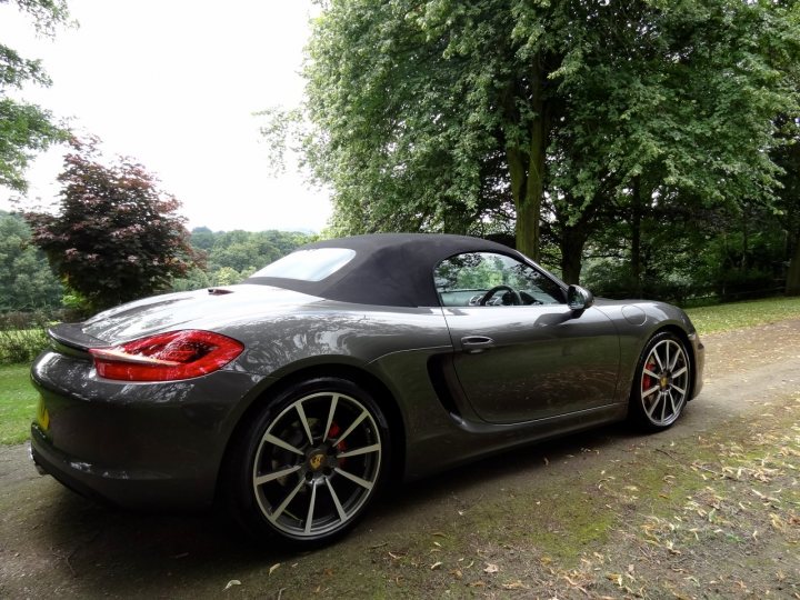 show us your toy - Page 69 - Porsche General - PistonHeads - This image showcases a sleek, dark grey Porsche sports car. The car is parked on what appears to be a gravel driveway, with a clear view of its rear and side profile. The Porsche sports a black convertible top, adding to its sporty aesthetic. The background is filled with lush green trees, creating a serene and contrasting backdrop for the car. The image does not contain any visible text or additional objects that would provide further context or information.