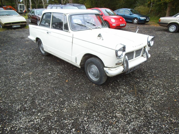 Classics left to die/rotting pics - Page 480 - Classic Cars and Yesterday's Heroes - PistonHeads - The image showcases a vintage white car parked on a gravel driveway. The car, characterized by its round headlights and black tires, appears to be in a condition of disrepair, with rust stains visible on its body. In the background, several other cars are parked, adding a sense of depth and the car's position amidst others. The scene gives a glimpse of a past era, encapsulated by the classic design of the older car.