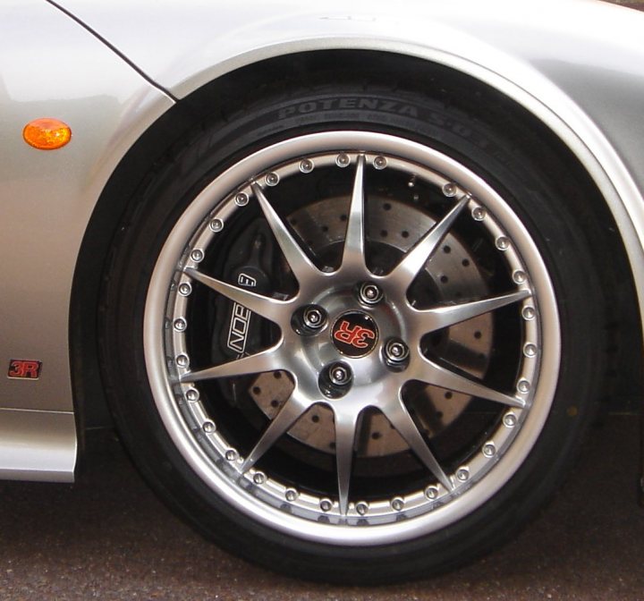 Powdercoat Noble M12GTO3R 12 spoke Wheels - Page 1 - Noble - PistonHeads - The image provides a close-up view of the front wheel of a silver car. The tire is positioned at an angle to the viewer, revealing a portion of its tread. The silver alloy rim is sharply contrasted against the metallic silver body of the car. The hubcap is visible and features a gold emblem in the center. The overall effect is a detailed look at the vehicle's wheel and the high-quality components it uses.