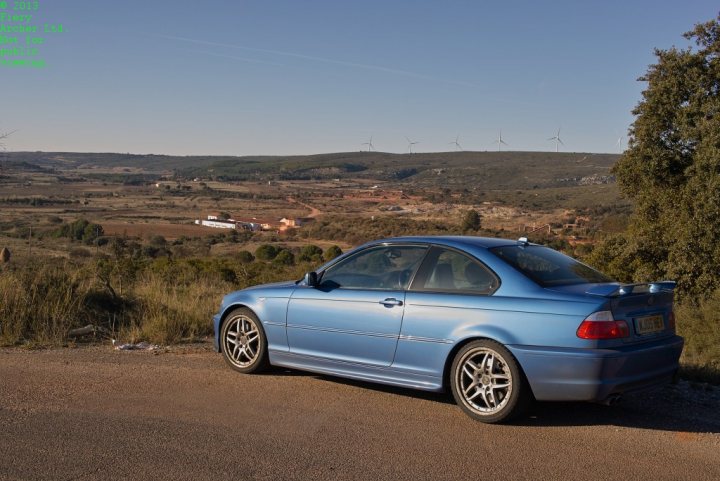 Anyone want some free photos of their cars? - Page 1 - East Anglia - PistonHeads - In the image, there's a striking blue car parked on the side of a road with a picturesque view in the background. The car, a BMW, sports a black hood and a black rear wing, contrasting with its vibrant blue body. The road seems lonely and serene, with a smattering of trees along the side and a lone building visible in the distance. In the far distance, several wind turbines stand against the horizon, adding a touch of industrial charm to the otherwise natural landscape. The sky above is a clear blue with only a few wispy clouds, suggesting a bright and sunny day. The overall scene is one of tranquility and openness.