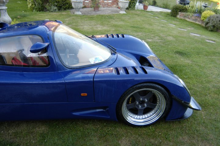 Wheel Pistonheads Louvres Arch - The image displays a blue sports car parked on a grassy yard. The car's sleek design includes a curved front and an aerodynamic side profile. It features a clear windshield and two large, inverted headlamps. The car is equipped with silver sports rims. There are a couple of potted plants visible in the background, adding a touch of greenery to the scene. The setting suggests a private garden environment.