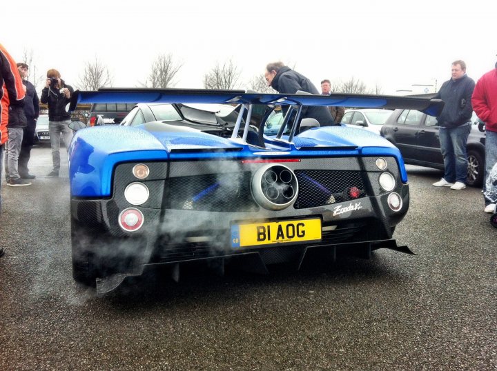 Saywell track day - Page 1 - Goodwood Events - PistonHeads - The image captures an outdoor setting where a striking blue single-seater sports car is parked. The car features a noticeable red stripe running along its roof and a black rear wing. It's equipped with clear headlights and a prominent black grille. Scattered around the car, there are several people who seem to be observing the vehicle, contributing to a casual, crowd-like atmosphere.