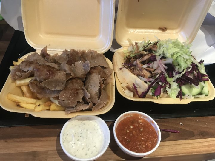 Dirty Takeaway Pictures Volume 3 - Page 333 - Food, Drink & Restaurants - PistonHeads - The image shows a table with two takeaway containers of food. One container has what appears to be a kebab or shish kebab, served with fries and salad on the side. The other container holds a dish that looks like a gyro or wrap, accompanied by a salad with cabbage, carrot, and lettuce. Next to each takeaway is a small condiment cup containing sauce, possibly tzatziki or another type of dip. The overall setting suggests a casual dining environment, likely a cafe or a similar establishment that offers quick meals.