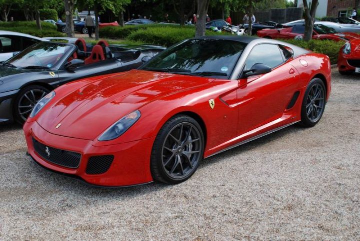 Pistonheads Supercar Sunday - This image showcases a close-up of a vibrant red sports car parked on a gravel surface in what appears to be a parking lot or outdoor exhibition area. The car is sleek and stylish, featuring a distinctive design and what looks like a television screen on its side, indicative of the sportscar's high-tech and performance-oriented nature. The background reveals other vehicles and people scattered about, suggesting a public event or gathering. There is a sense of motion and dynamism conveyed by the bright red color of the sports car, contrasting with the more subdued colors of the other vehicles. Overall, the image captures a moment of automotive splendor amidst a lively setting.