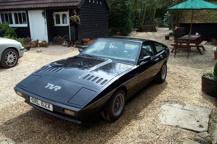 Pistonheads Favourite Wedge - The image shows a black sports car parked on a gravel driveway. The car is positioned at an angle that partially obscures its front right side. The vehicle has a distinctive C-shaped vent on its hood, reminiscent of classic sports cars. It also features a front lip, which adds to its aggressive and sporty design. The car is parked in front of a house with a black pitched roof, partially obscured by a large, attractive tree. The setting seems to be a residential area, possibly a garden or a driveway of a private property. The photograph conveys a sense of personal taste and interests in automobiles.